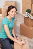 Marking a carton box. Top view of cheerful young woman marking a cardboard box and looking at camera while more boxes laying on background photo