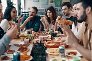 tener hambre. grupo de jóvenes con ropa informal comiendo pizza y sonriendo mientras cenan en el interior foto