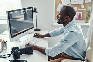 apuesto joven africano usando computadora y sonriendo mientras trabaja en la oficina moderna foto