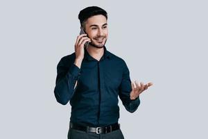 Discussing new projects. Handsome young smiling man talking on smart phone and gesturing while standing against grey background photo