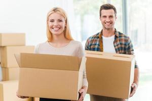 Moving to a new house together. Cheerful young couple holding cardboard boxes while other carton boxes laying on background photo