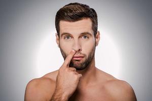 Digging for gold. Portrait of handsome young shirtless man picking his nose while standing against grey background photo