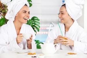 Spending time at spa. Two beautiful young women in bathrobe drinking tea and talking to each other while sitting in front of swimming pool photo