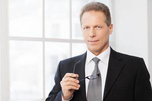 Smart and confident. Portrait of confident mature man in formalwear holding glasses and smiling at camera while standing near window photo