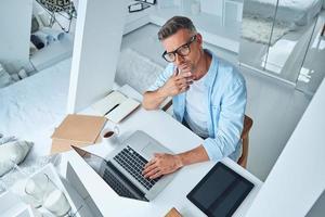 Top view of confident mature man working on laptop and looking at camera while sitting at the office desk photo