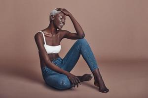 belleza inolvidable. atractiva joven africana sonriendo mientras se sienta en un fondo marrón foto