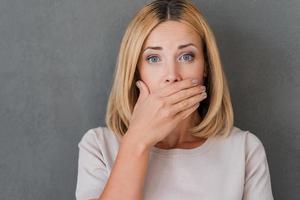 Shocking news. Surprised mature woman covering mouth with hand and staring at camera while standing against grey background photo