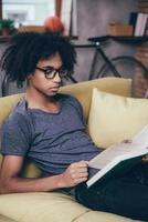 Reading book at home. Side view of young African man reading book and wearing glasses while sitting on the couch at home photo