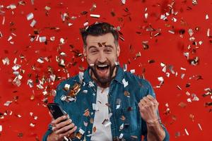 joven feliz con ropa informal sosteniendo un teléfono inteligente y animando mientras está de pie contra un fondo rojo con confetti volando alrededor foto