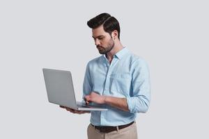 Concentrated at work.  Good looking young man working on his laptop while standing against grey background photo