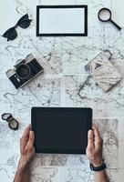 Ready to travel. Close up top view of man using digital tablet with sunglasses, photo camera, compass, magnifying glass and passport lying on map around