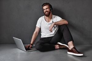 Happy man.  Handsome young man listening music and using computer while sitting on the floor against grey background photo