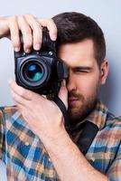 Serious photographer. Portrait of confident young man in shirt holding camera in front of his face while standing against grey background photo