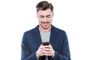 Chatting with friend. Handsome young man typing message on mobile phone and smiling while standing against white background photo