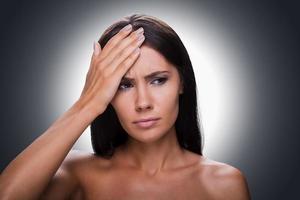 Feeling awful headache. Portrait of frustrated young shirtless woman looking away and touching forehead with hand while standing against grey background photo