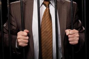 White collar crime. Cropped image of man in formalwear standing behind a prison cell while isolated on black background photo