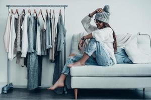 Choosing the right option.  Thoughtful young woman in casual wear adjusting her hat and looking away while sitting on the couch at home near her clothes hanging on the racks photo