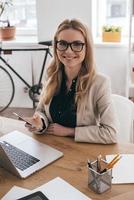 Beautiful business lady. Gorgeous young woman in smart casual wear holding smart phone and looking at camera while sitting at her working place in creative office photo