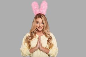 Please Attractive young woman in pink bunny ears keeping hands clasped and looking at camera while standing against grey background photo