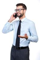 Staying in touch with his colleagues. Confident young handsome man in shirt and tie talking on mobile phone and smiling while standing against white background photo