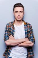 CHarming handsome. Handsome young man in shirt looking at camera and keeping arms crossed while standing against grey background photo