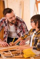 Working together. Smiling young male carpenter and his son looking at each other while working in workshop photo