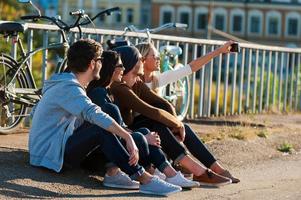 tiempo para selfie. grupo de jóvenes sonrientes que se unen entre sí y se hacen selfie por teléfono inteligente mientras se sientan al aire libre junto con bicicletas en el fondo foto