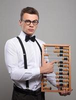 Financial advisor. Serious young man in bow tie and suspenders holding abacus and pointing it while standing against grey background photo