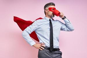 hombre confiado en camisa y corbata usando capa de superhéroe y bebiendo bebida caliente contra fondo rosa foto