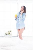 Calling to say good morning. Beautiful young woman in blue shirt talking on the phone and holding a glass of juice while standing near the bed photo