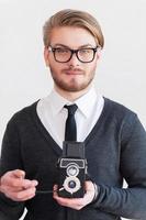 fotógrafo de estilo antiguo. un joven apuesto con anteojos sosteniendo una cámara retro mientras se enfrenta a un fondo gris foto