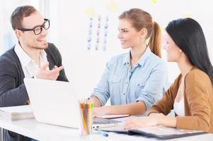 Brainstorming. Three young colleagues sitting together at their working place while woman telling something and gesturing photo