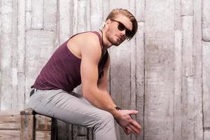 Cool and trendy. Side view of handsome young man in sunglasses sitting on the wooden box and looking away photo