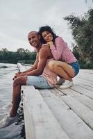 dos corazones llenos de amor. feliz pareja joven abrazándose y sonriendo mientras se sienta en el muelle cerca del lago foto