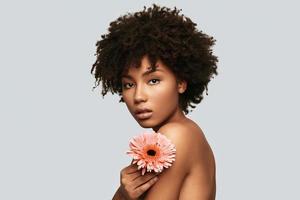 Irresistible girl. Attractive young African woman looking at camera and holding a flower while standing against grey background photo