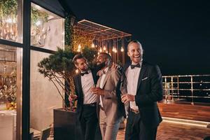 Three happy men in suits holding glasses with whiskey while spending time on party photo