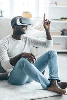 Pointing somewhere. Handsome young African man in smart casual clothes pointing somewhere and smiling while sitting on the carpet at home photo