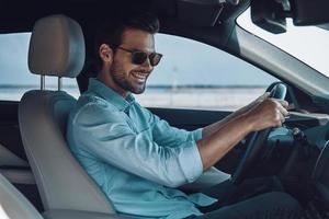 feliz de conducir un coche nuevo. un joven apuesto con ropa informal inteligente sonriendo mientras conduce un coche de estado foto