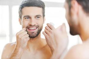 Cleaning teeth with dental floss. Rear view of handsome young shirtless man cleaning his teeth with dental floss and smiling while standing in front of the mirror photo