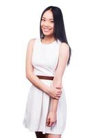 Charming beauty. Beautiful young Asian woman in pretty dress looking at camera and smiling while standing against white background photo