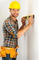 Everything should be perfect. Cheerful young repairman making measurements on the wall by level and looking at camera while standing indoors photo