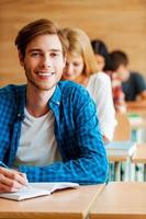 He is a top student. Happy young man writing in note pad and looking at camera while his classmates sitting behind him at their desks in the classroom photo