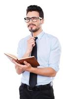 Just inspired. Thoughtful young man in shirt and tie making notes in his pad and smiling while standing against white background photo