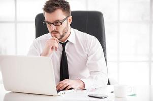 empresario en el trabajo. guapo joven barba hombre en camisa y corbata trabajando en la computadora portátil mientras está sentado en su lugar de trabajo foto