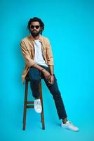 Full length of charming young African man in casual wear looking at camera while sitting on stool against blue background photo