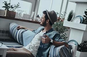 Enjoying fresh coffee. Thoughtful young man holding a cup and looking outside while sitting on the sofa photo