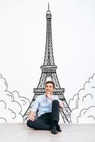 Dreaming about Paris. Young handsome man keeping hand on chin and looking away with smile while sitting on the floor with illustration of Eiffel Tower at the background photo