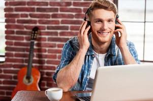 disfrutando de la musica un joven apuesto con auriculares trabajando en una laptop y sonriendo mientras la guitarra acústica está en el fondo foto