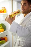 Midnight eating. Surprised young man in bathrobe standing near the open fridge and eating photo