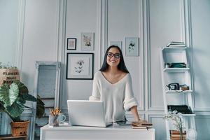 Young and successful. Beautiful young woman looking at camera and smiling while standing in home office photo